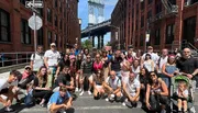 A diverse group of people pose for a photo on a sunny day with the iconic Manhattan Bridge in the background.