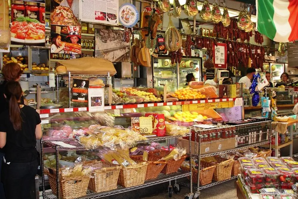 A bustling deli features an array of goods from hanging meats to fresh produce with customers and a worker visible amidst the colorful display