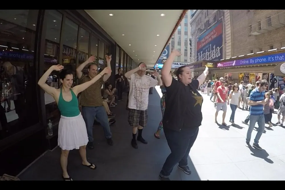 A group of people are dancing on a busy sidewalk in front of a store with onlookers passing by