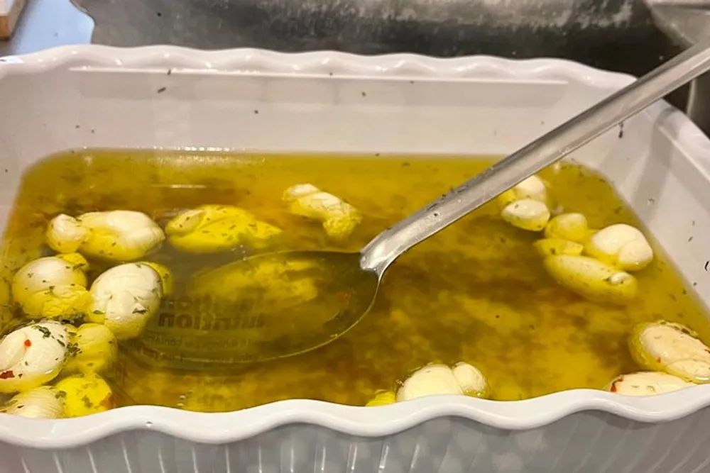 A white plastic container holds a clear yellow broth with herbs and numerous peeled garlic cloves alongside a silver soup ladle