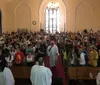 The image shows a congregation of people inside a church standing as they face the aisle with a clergy member walking down the center