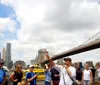 A tour guide is gesturing and explaining something to a group of attentive tourists with the Brooklyn Bridge and the Manhattan skyline in the background