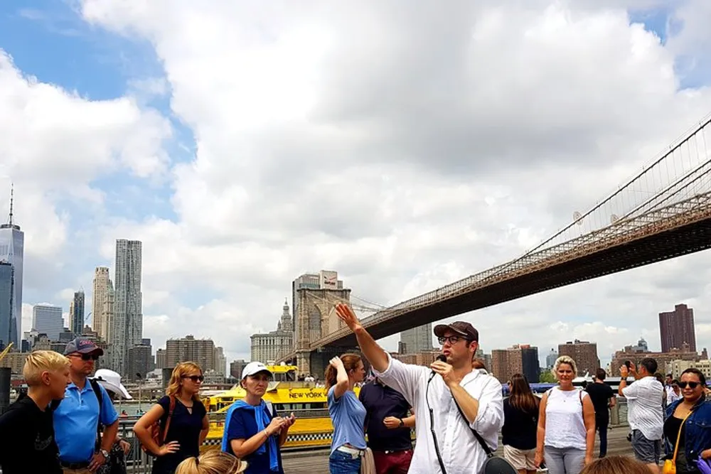 A tour guide is gesturing and explaining something to a group of attentive tourists with the Brooklyn Bridge and the Manhattan skyline in the background