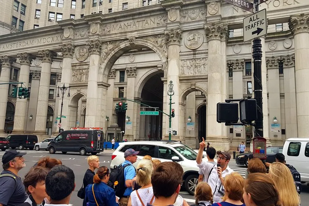 A group of people on a city tour are listening to a guide who is pointing towards an ornate building with the inscription MANHATTAN above the archway
