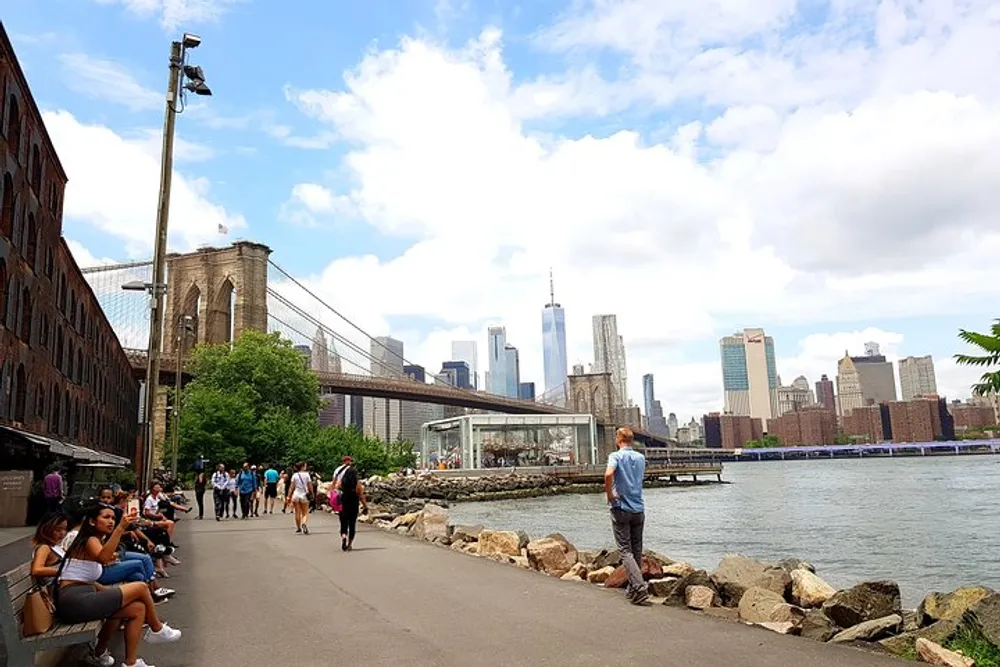 This image shows a lively waterfront walkway with pedestrians the Brooklyn Bridge to the left and the Manhattan skyline including the One World Trade Center in the background under a partly cloudy sky