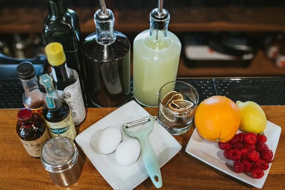 The image displays various cocktail ingredients and tools including citrus fruits berries eggs bitters syrups and a juicer arranged on a bar counter