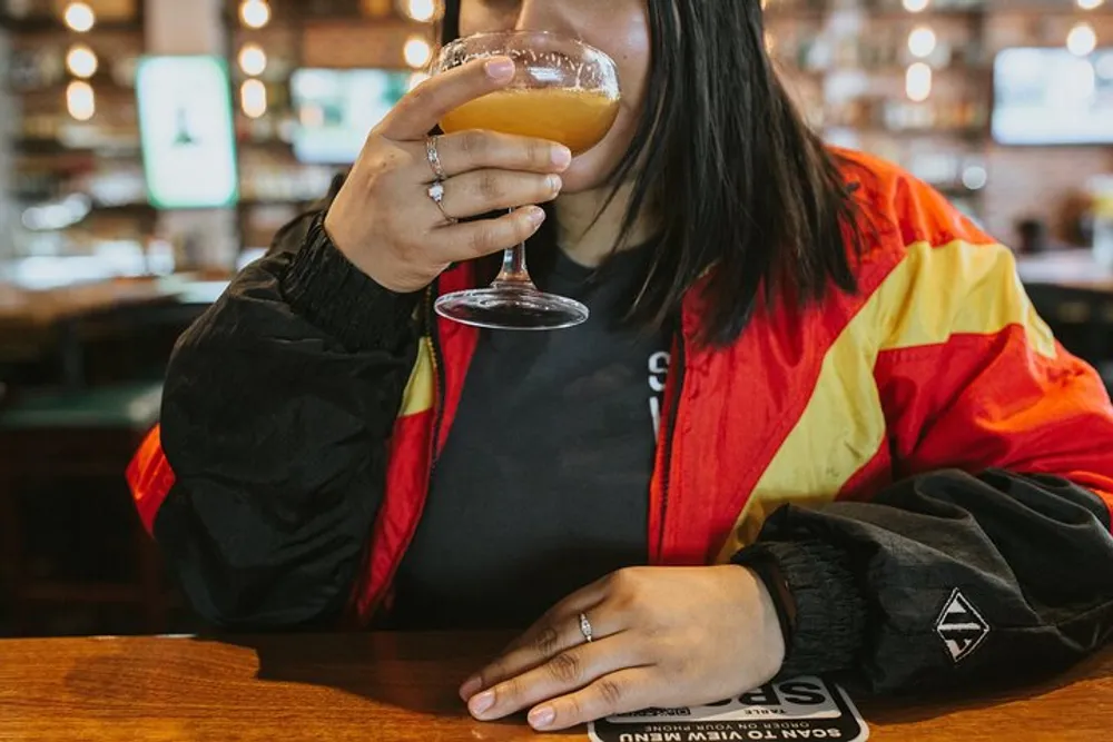 A person in a colorful jacket is sipping a beverage at a bar or pub with the setting featuring warm lighting and blurred bar shelves in the background