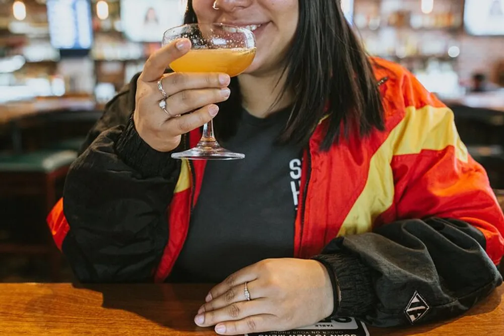 A smiling person in a colorful jacket is raising a glass of drink to their mouth at a bar or restaurant