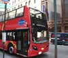 A red double-decker sightseeing tour bus is parked on a city street with cars passing by and urban buildings in the background