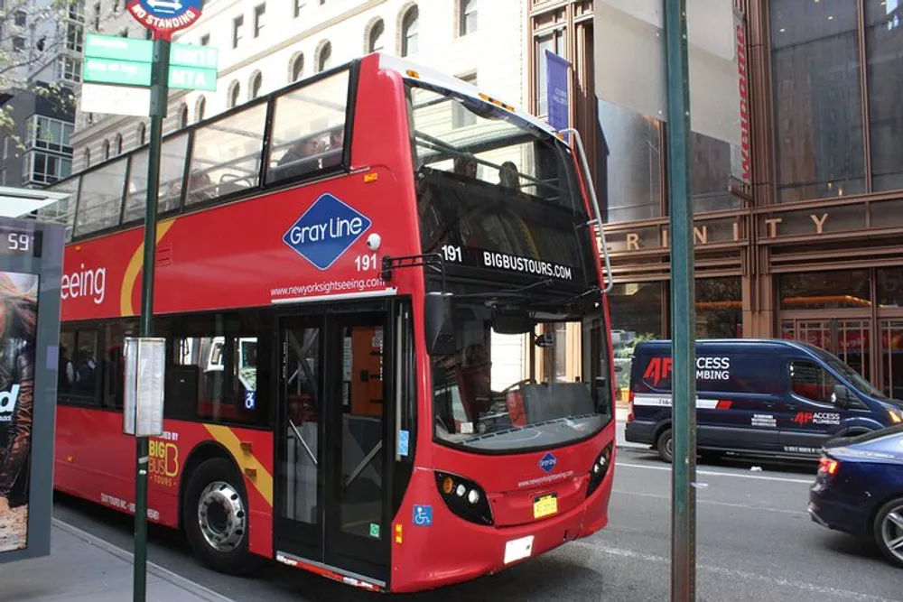 A red double-decker sightseeing tour bus is parked on a city street with cars passing by and urban buildings in the background