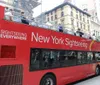 A red double-decker sightseeing tour bus is parked on a city street with cars passing by and urban buildings in the background