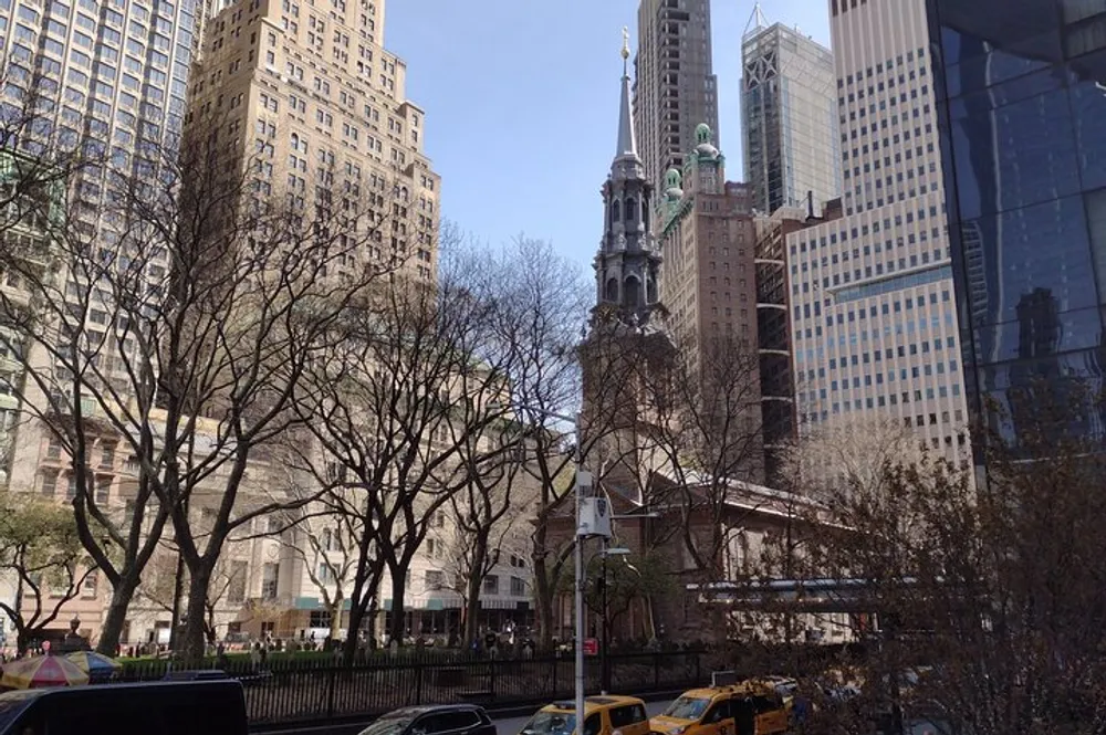 This is a street-level view of a bustling urban environment with a mix of modern and historic architecture including a church with green-topped spires surrounded by leafless trees and yellow taxis in the foreground