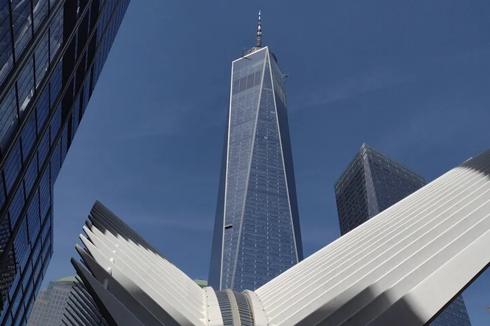 The image shows the One World Trade Center tower rising majestically behind the distinctive white wing-like structure of the Oculus pavilion under a blue sky