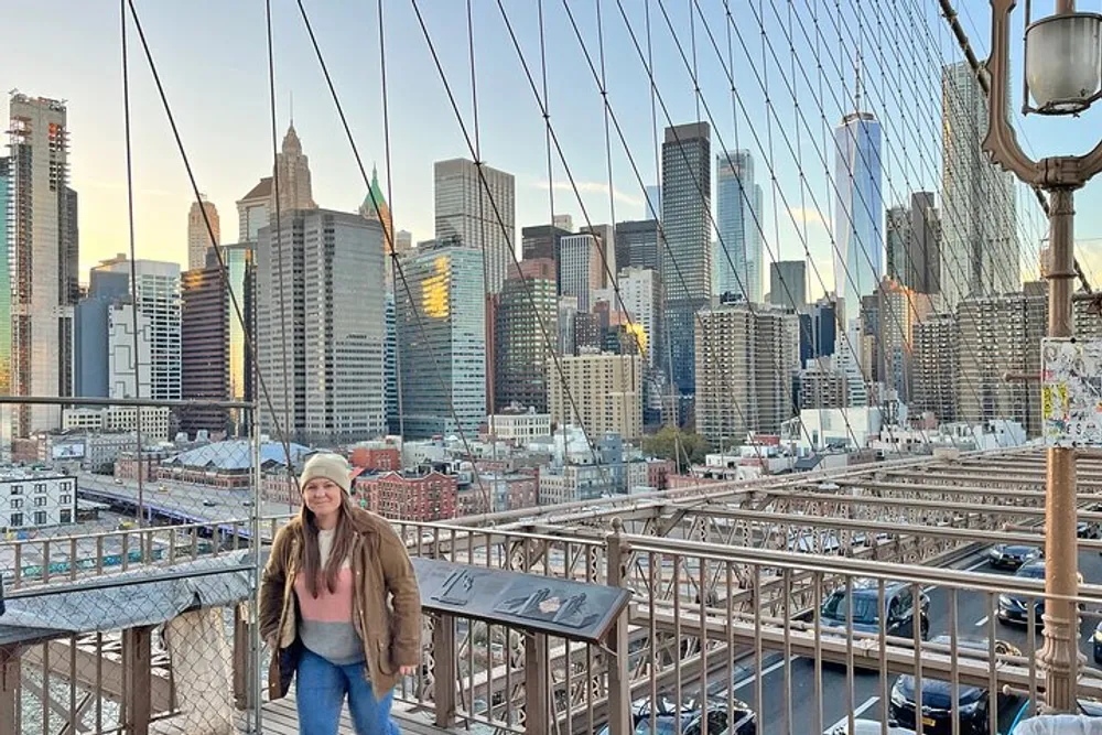 A person is posing for a photo on what appears to be the Brooklyn Bridge with a backdrop of the Manhattan skyline during the daytime