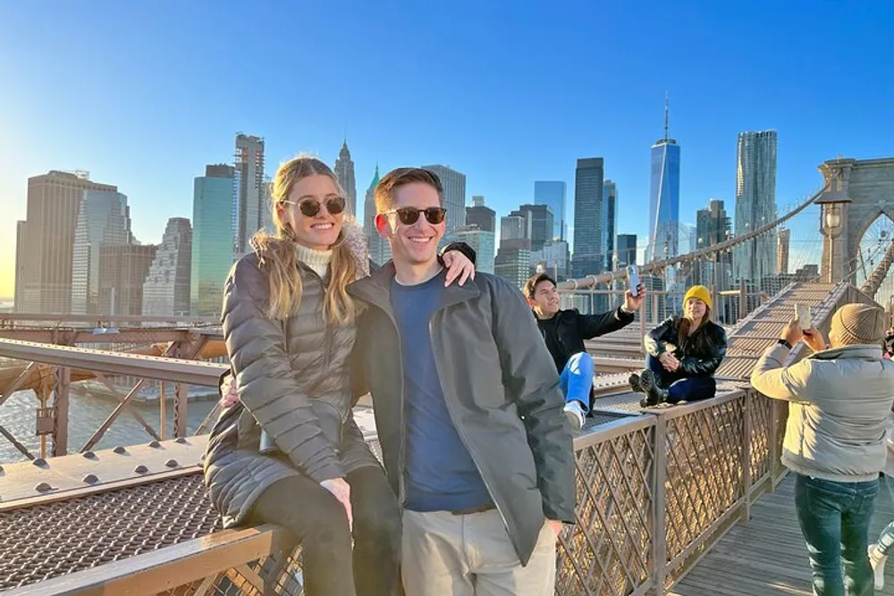 A smiling pair poses for a photo on a sunny day with the New York City skyline and the Brooklyn Bridge in the background