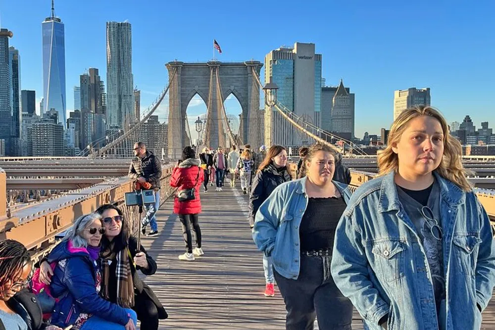 People are walking across the Brooklyn Bridge with the New York City skyline in the background on a sunny day