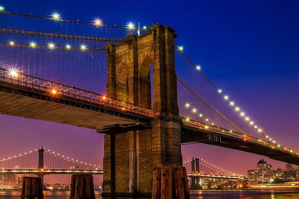 A nighttime view of the Brooklyn Bridge in New York City with the Manhattan Bridge in the background both illuminated against the evening sky