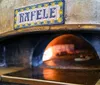This image shows a close-up view of a traditional wood-fired pizza oven with a glowing fire at the back and the name Raffaele on a decorative ceramic tile above the entrance