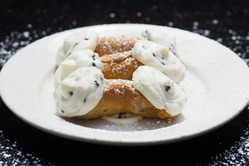 The image shows a dessert likely a cannoli filled with cream and dotted with chocolate chips sprinkled with powdered sugar and served on a white plate