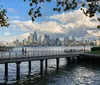The image features a scenic view of a city skyline beyond a water body captured from a shaded park area with people walking on a pier