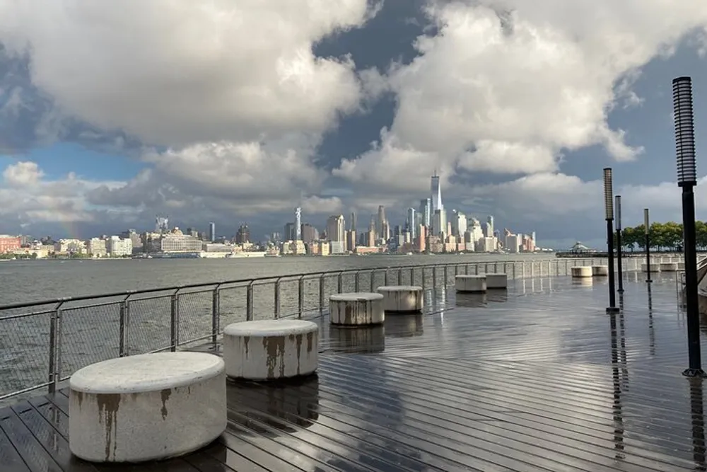 The image shows a waterfront view of a city skyline with high-rise buildings under a dramatic sky with a partial rainbow taken from a wet wooden pier with seating bollards