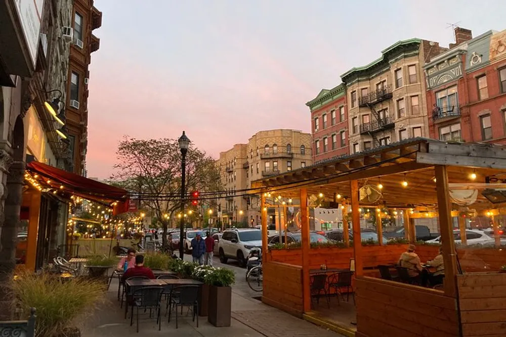An urban street scene at dusk with outdoor dining string lights and vintage buildings under a pink-tinged sky