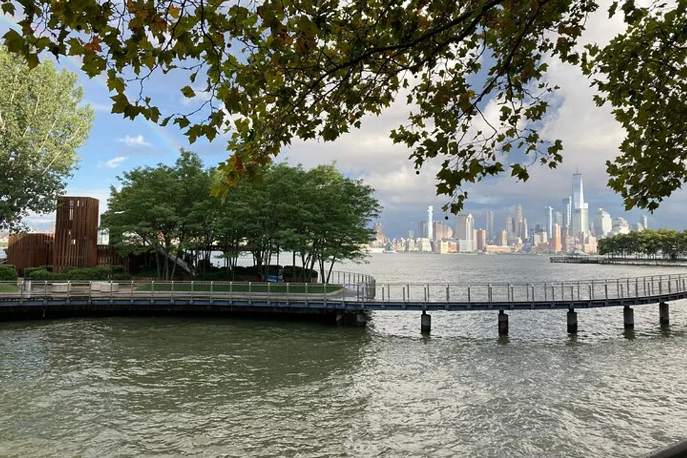 This image features a waterfront park with a wooden pier extending over the water framed by overhanging tree branches with a backdrop of a distant city skyline under a cloudy sky
