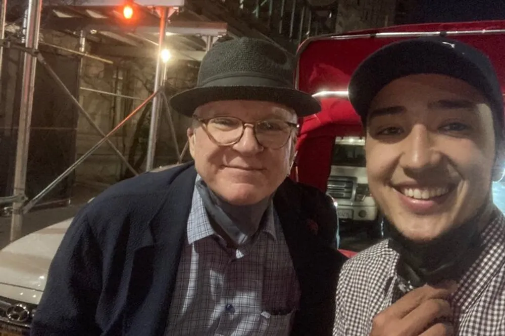Two smiling men one older wearing a fedora and the other younger with a cap take a selfie together at night against an urban backdrop with construction scaffolding