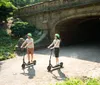 Two people wearing helmets are enjoying a ride on electric scooters near a historical archway in a park setting