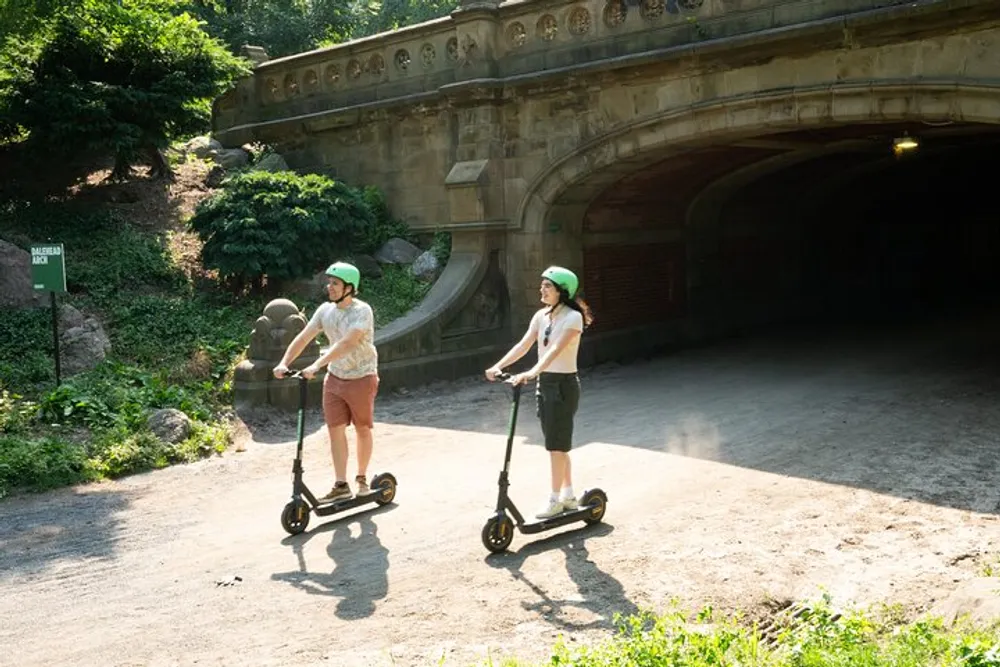 Two people wearing helmets are enjoying a ride on electric scooters near a historical archway in a park setting