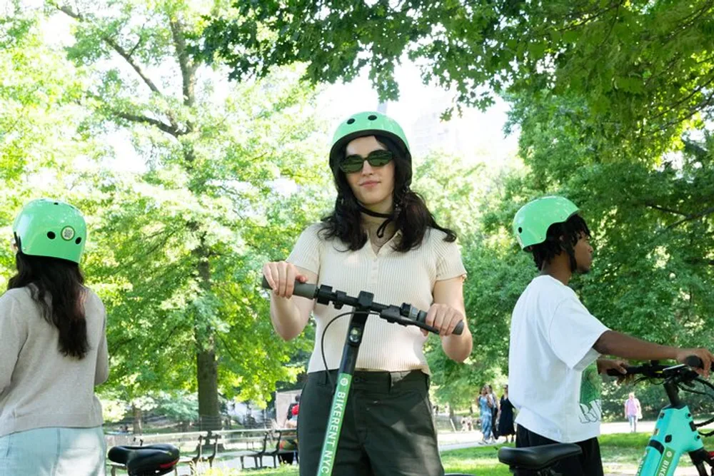 Three individuals wearing green helmets are in a park with the one in the foreground grasping the handlebars of an electric scooter while the others may be preparing to ride theirs