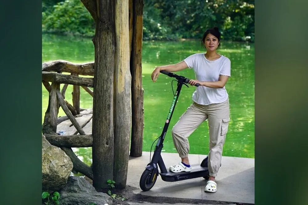 A person is standing next to an electric scooter in a park-like setting with greenery in the background