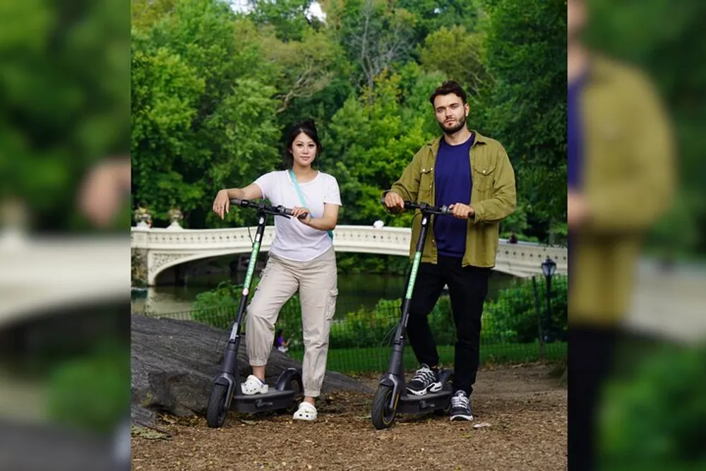 Two people are standing with electric scooters in a park with a bridge in the background