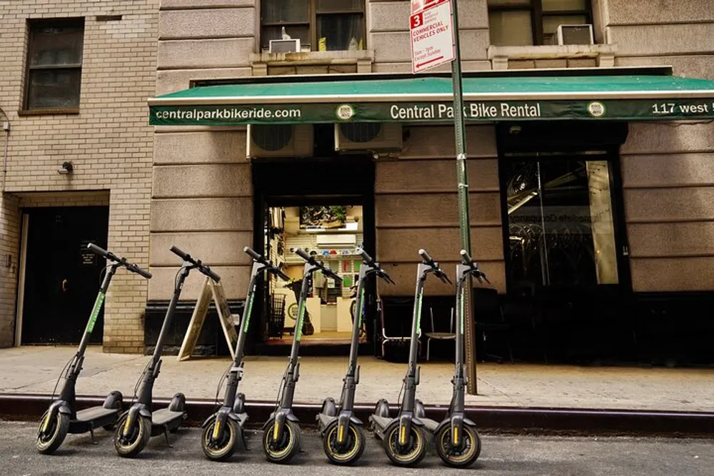 A row of electric scooters is parked in front of a bike rental shop near Central Park