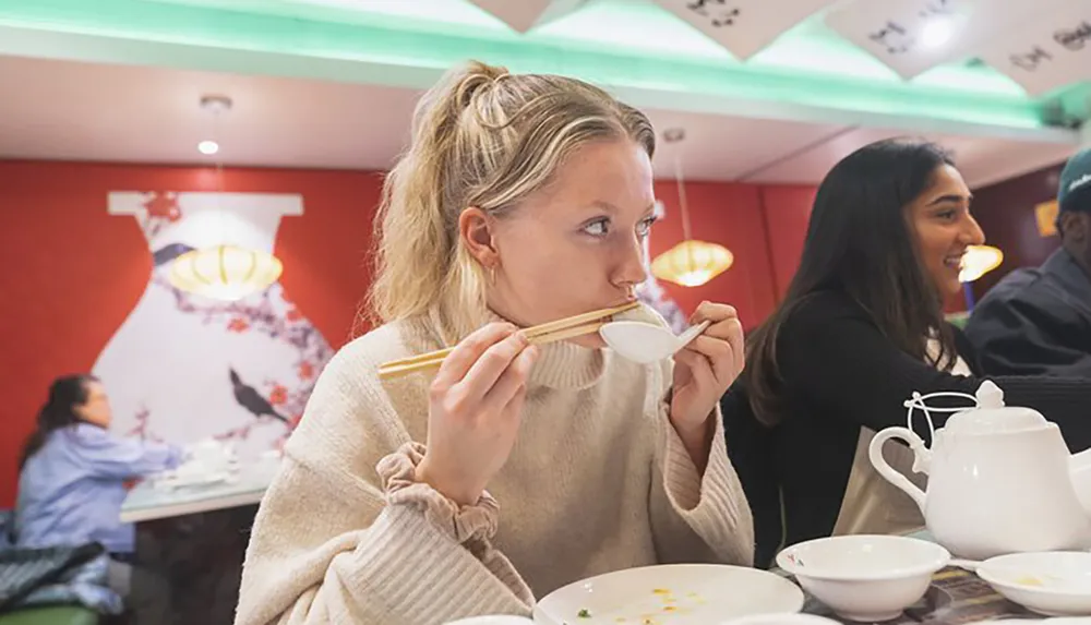 A person is enjoying a meal with chopsticks and sipping soup from a spoon in a vibrant restaurant setting