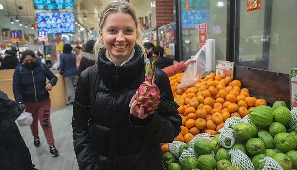 A smiling person is holding a dragon fruit in a bustling indoor market with a variety of fruits on display