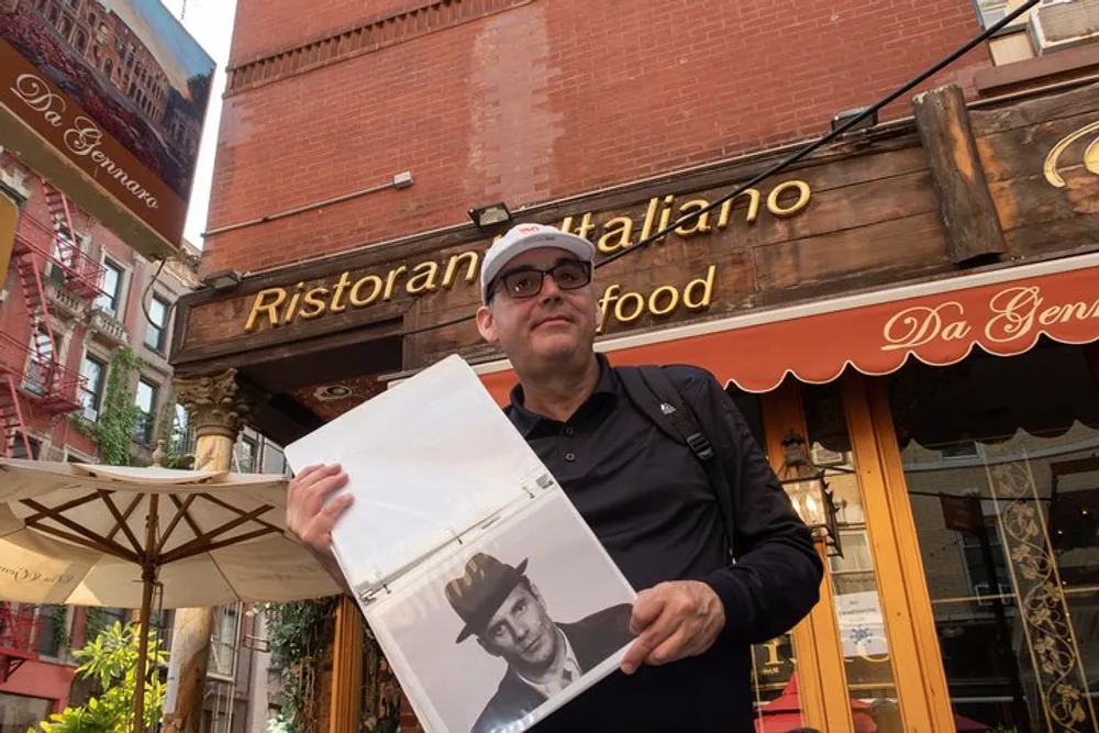 A man is standing in front of an Italian restaurant holding up a clear plastic sleeve with a black and white photograph inside