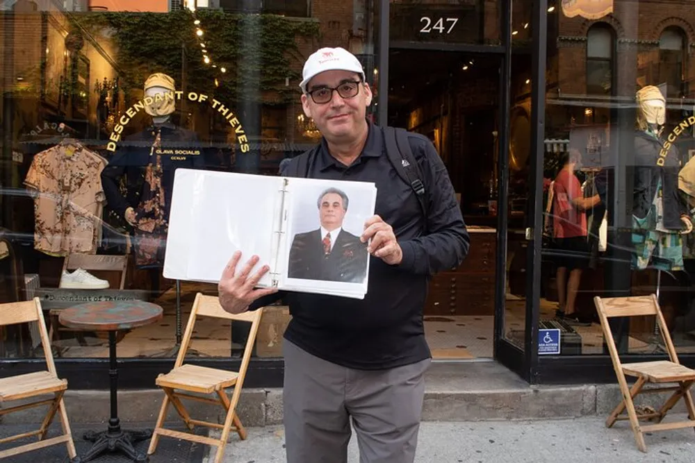 A man in glasses and a cap is standing on a sidewalk showing a page from a book with a portrait of a man in a suit