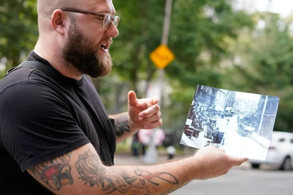 A tattooed man with glasses is animatedly talking while holding a black-and-white photograph of a city street