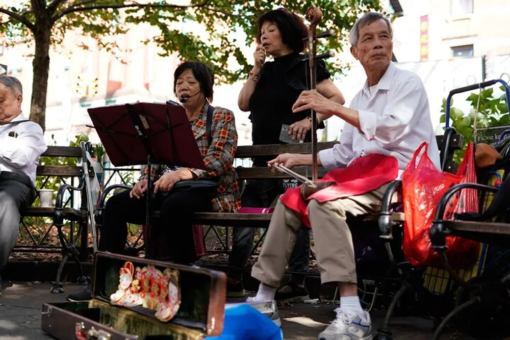 A group of elderly musicians are performing outdoors on a park bench with one woman singing and another playing a traditional stringed instrument surrounded by personal belongings and an open instrument case possibly for tips