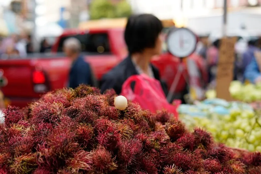 A large pile of rambutan fruit is in the foreground with a blurry marketplace and people in the background