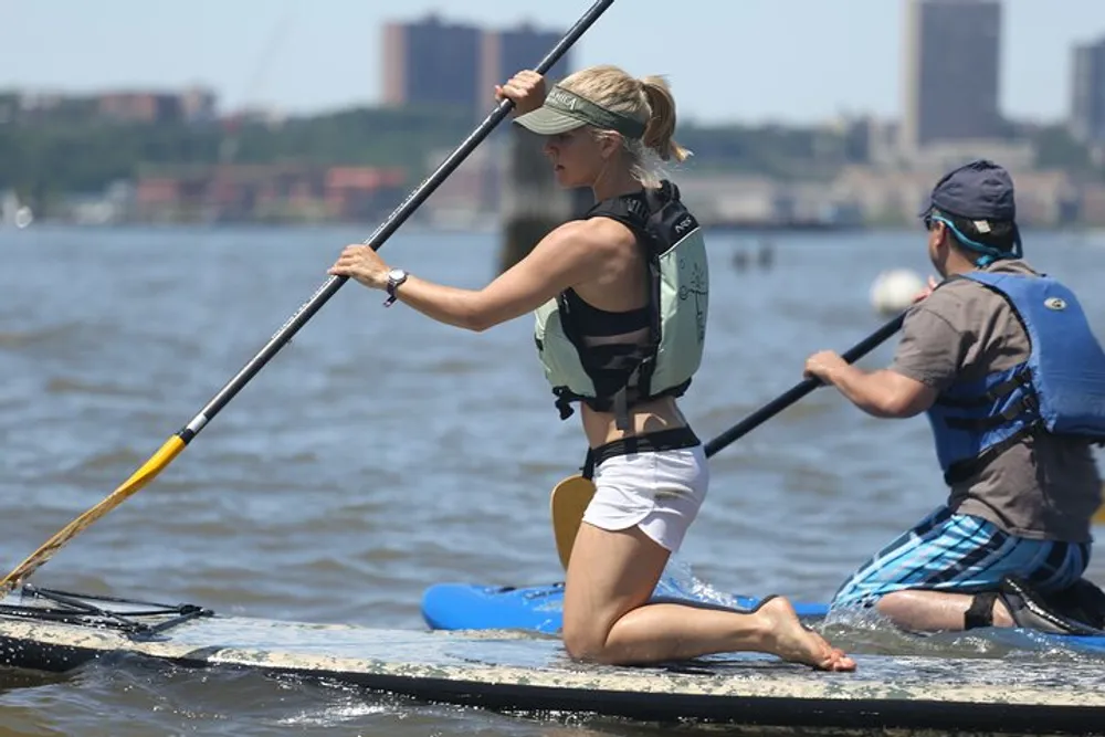 A woman and a man are paddleboarding on a body of water on a sunny day wearing life jackets and using paddles