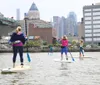 A person is stand-up paddleboarding on a river in front of a cityscape during daytime