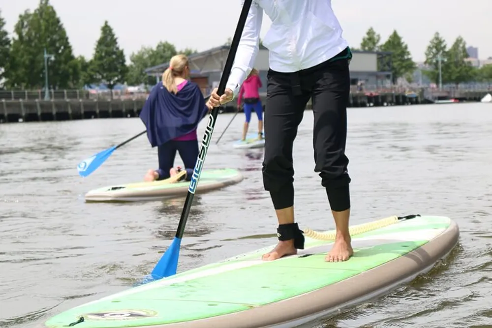 A person is stand-up paddleboarding on a calm body of water holding a paddle with others doing the same in the background