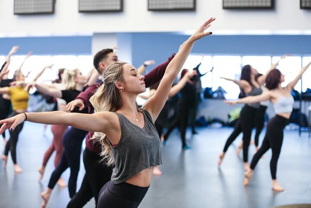 A group of dancers practices in a studio with a young woman in the foreground striking a graceful pose with her arms extended