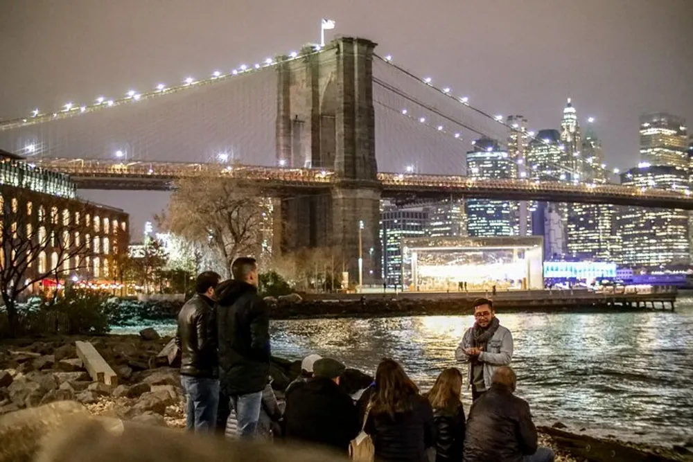 A group of people gather by the waters edge at night enjoying the view of the illuminated Brooklyn Bridge and the Manhattan skyline behind it
