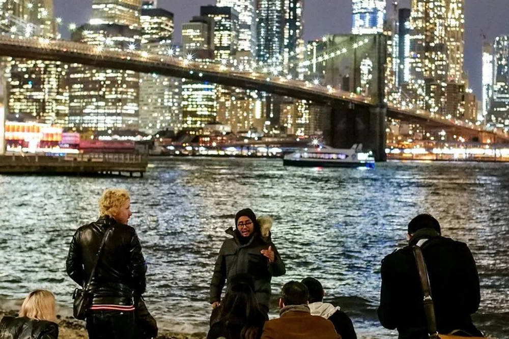 A group of people is gathered by a riverside at night with a brightly lit city skyline and a bridge in the background