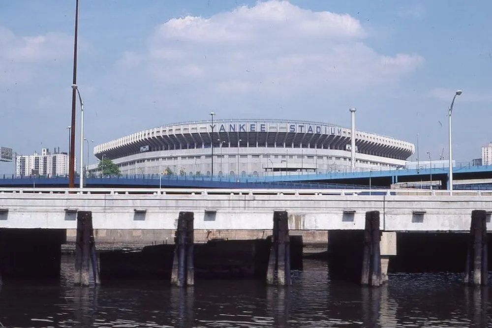 The image shows the exterior of the historic Yankee Stadium as viewed across water with a concrete pier in the foreground