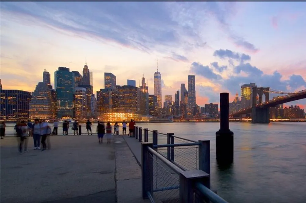 A group of people enjoy a sunset view of the Manhattan skyline and Brooklyn Bridge from a waterfront promenade