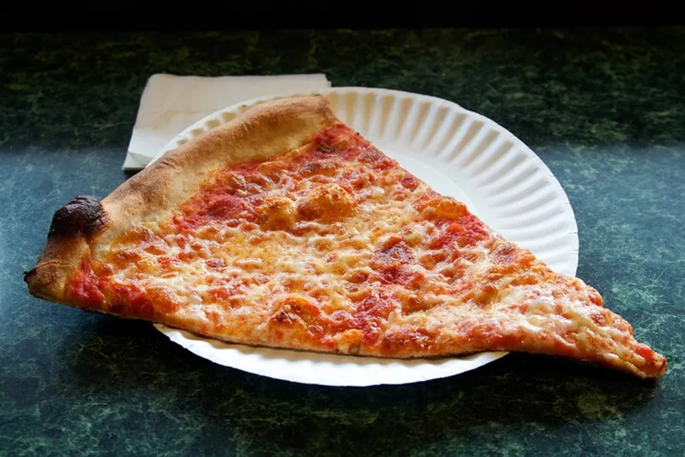 A single slice of cheese pizza is placed on a white paper plate on a greenish-black textured table with a wooden stirrer to the side